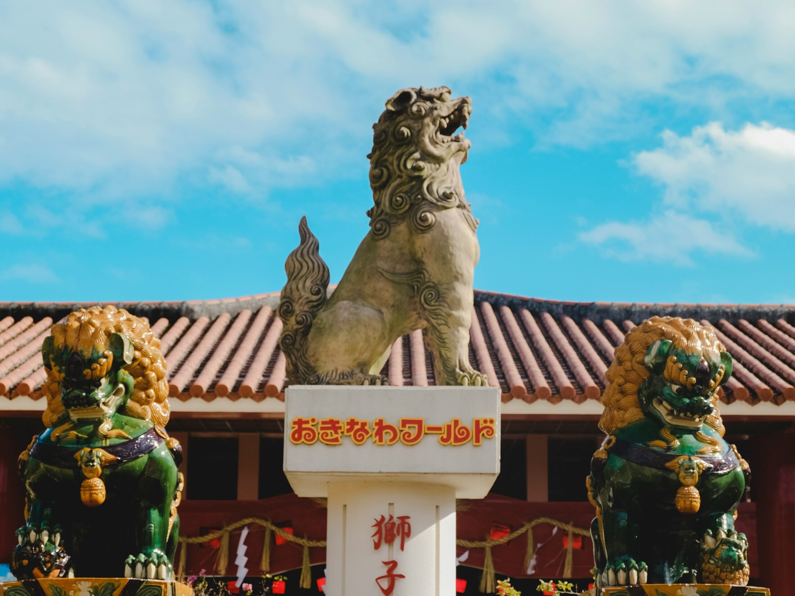 OKI-LIST-01 · a shisa on a red tile roof, seen from the street below. 4:3, 1400×1050