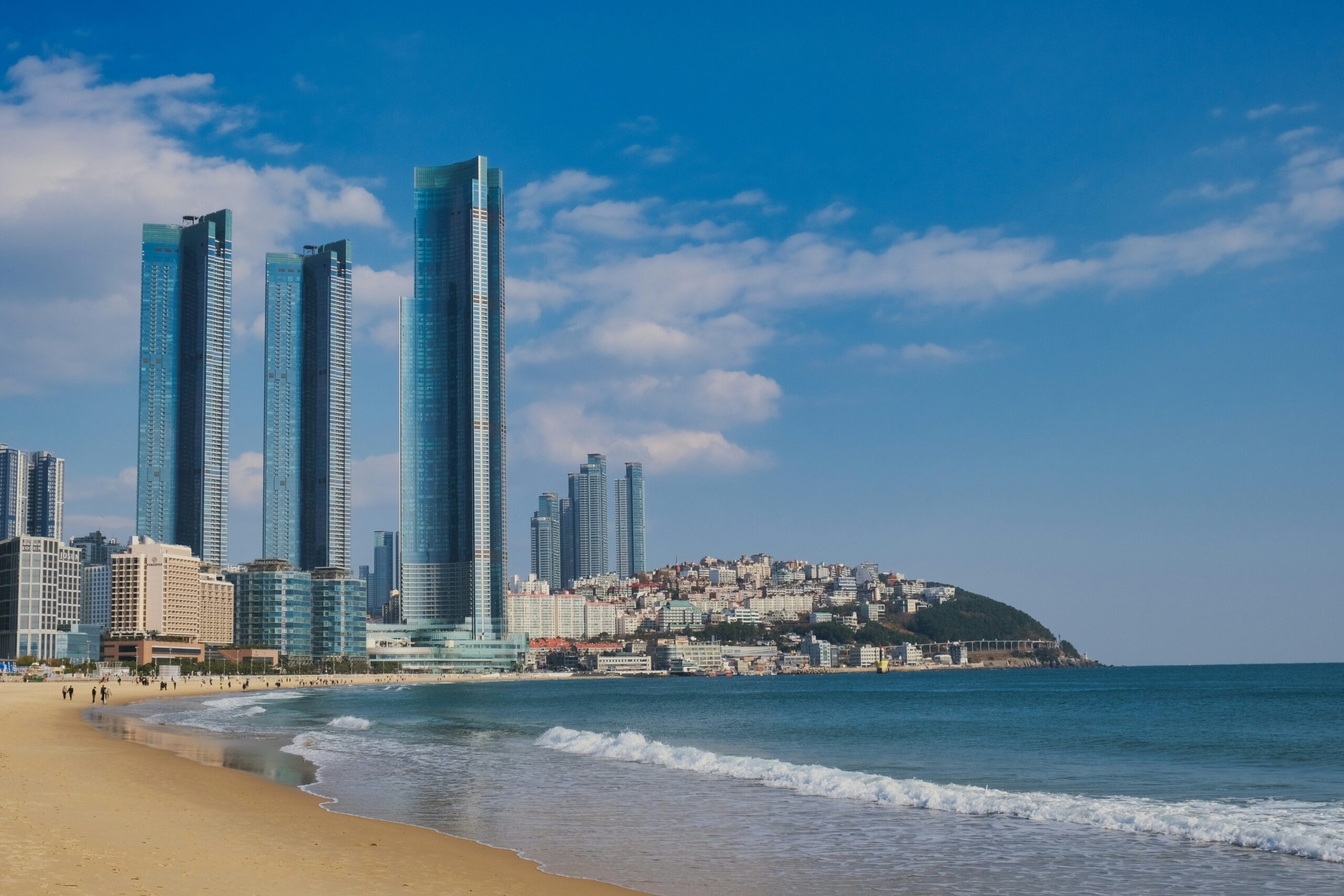 BS-COVER-01 · everyday Busan where city and sea coexist — hillside housing above the harbour edge, or a working waterfront street. No aerial skyline, no resort, no tourism-board beach. 3:2, 1800×1200