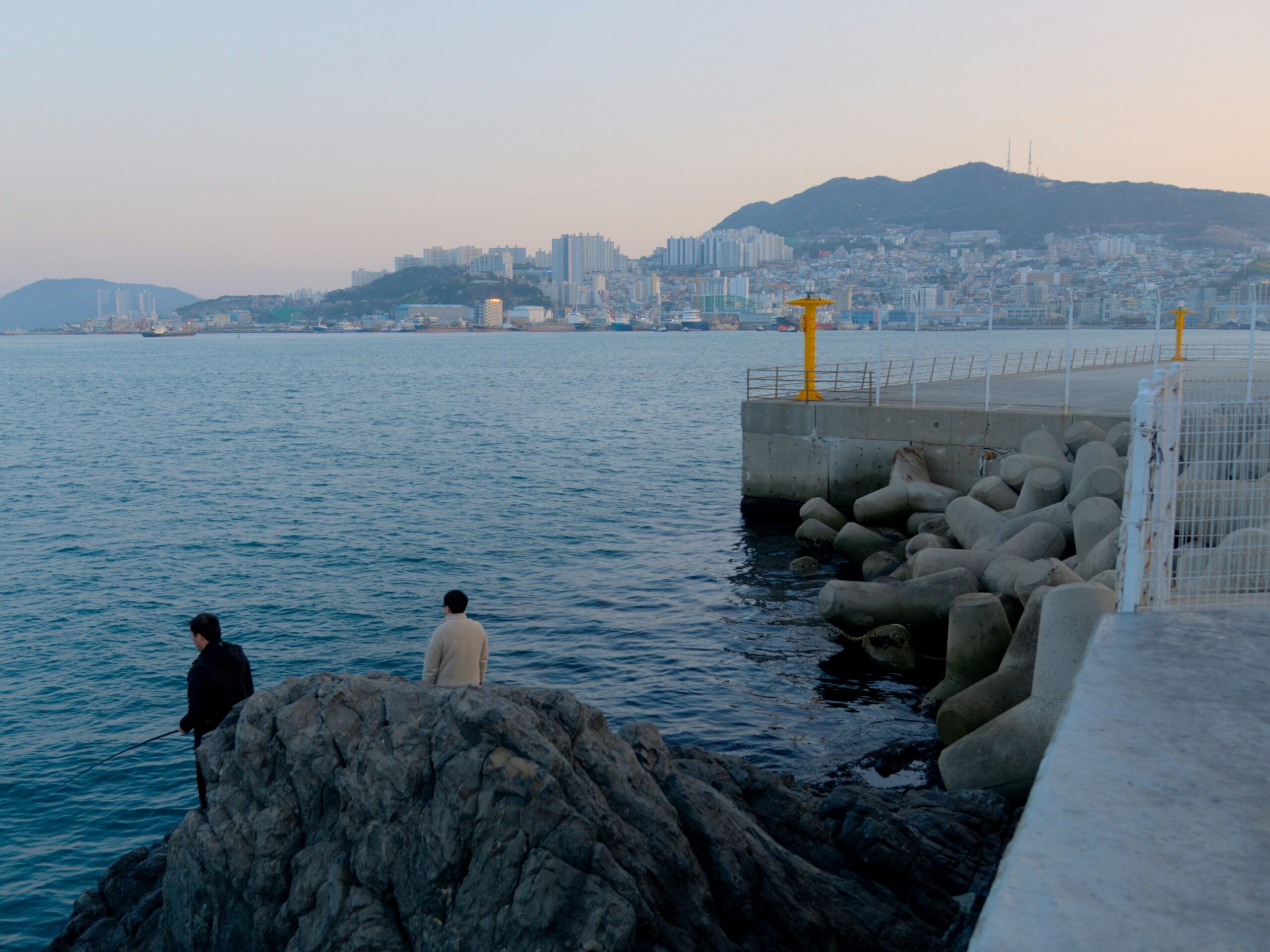BS-COAST-02 · local seaside — residents walking or fishing on a plain stretch of shoreline, late light. 3:2, 1800×1200
