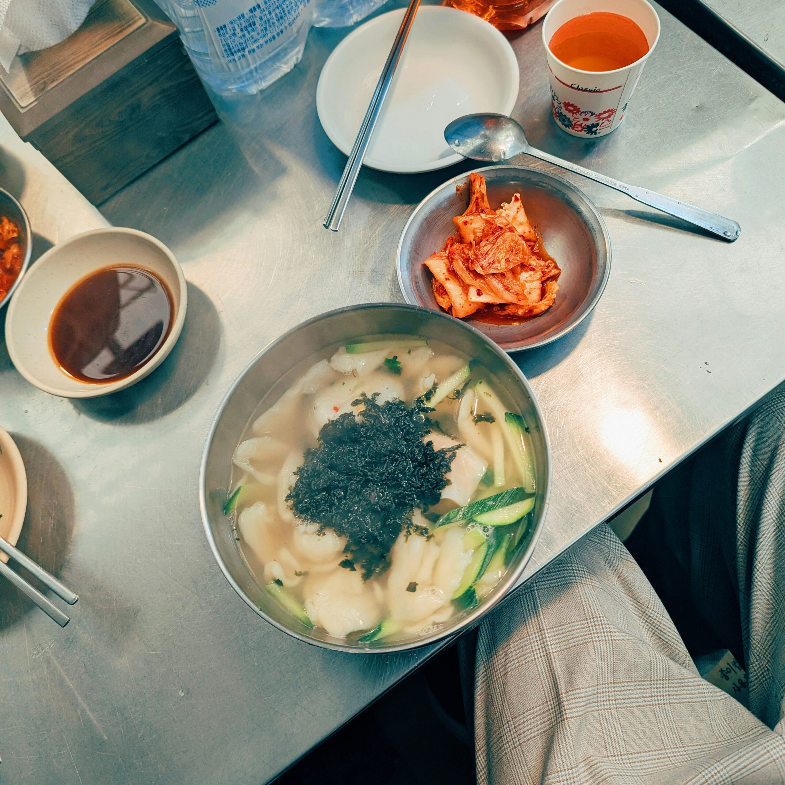 BS-EAT-01 · market breakfast — a bowl eaten standing up, steam, plastic stool. 1:1, 1000×1000