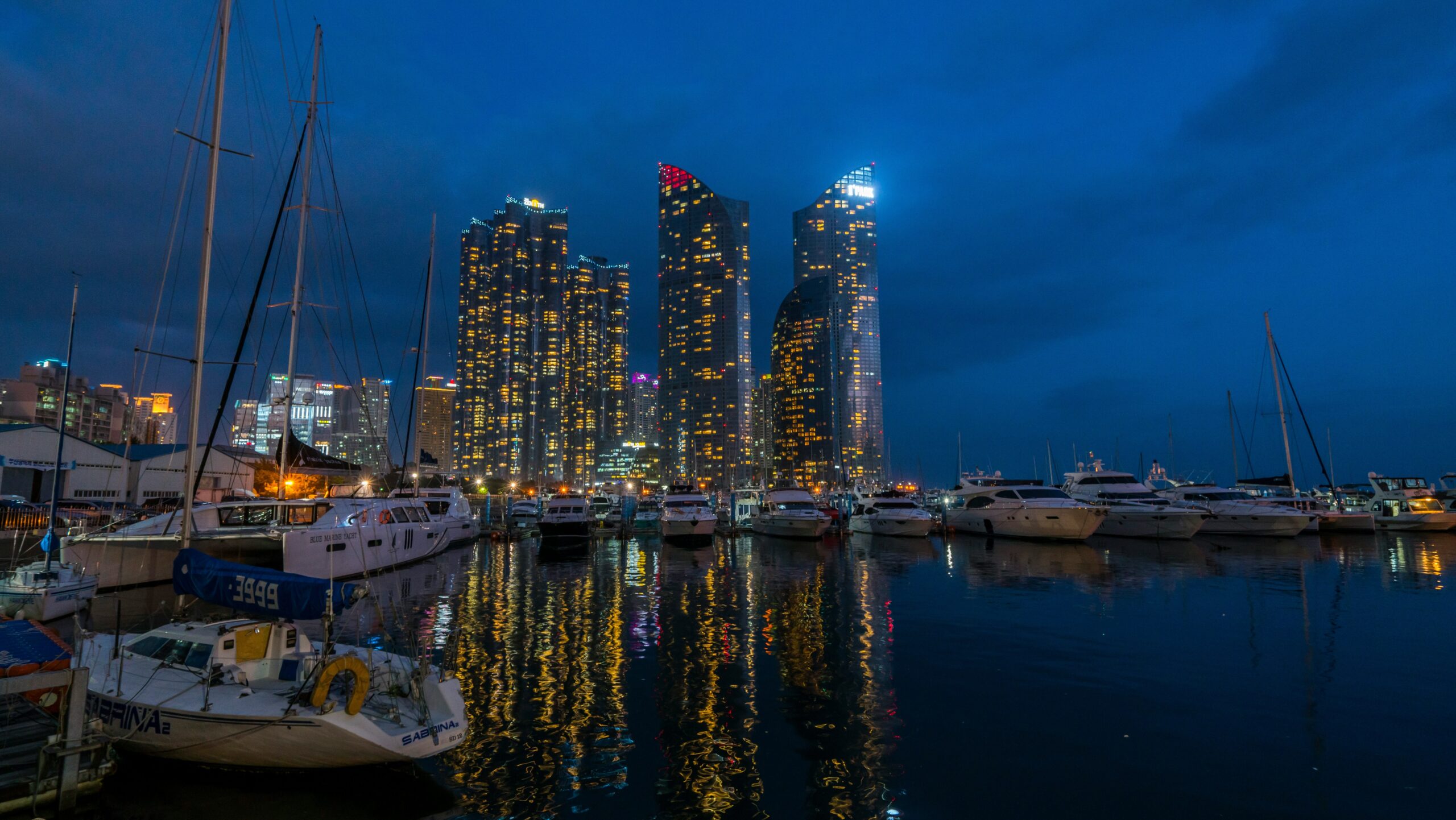 BS-NIGHT-01 · city lights along the bay reflected on water, believable exposure, no neon clichés. 16:9, 1920×1080
